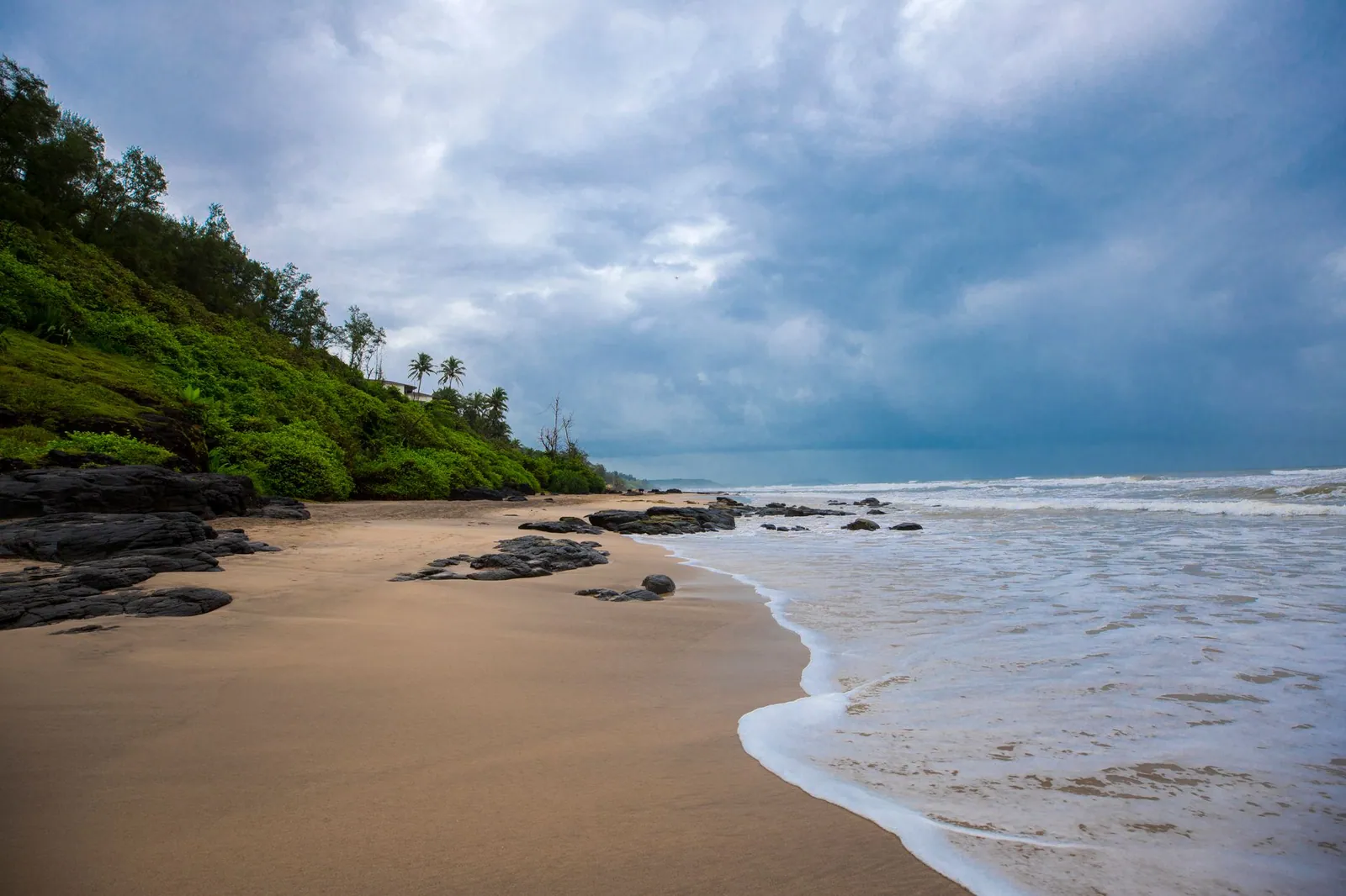 Explore this tranquil Indian beach with lush greenery and a dramatic cloudy sky.