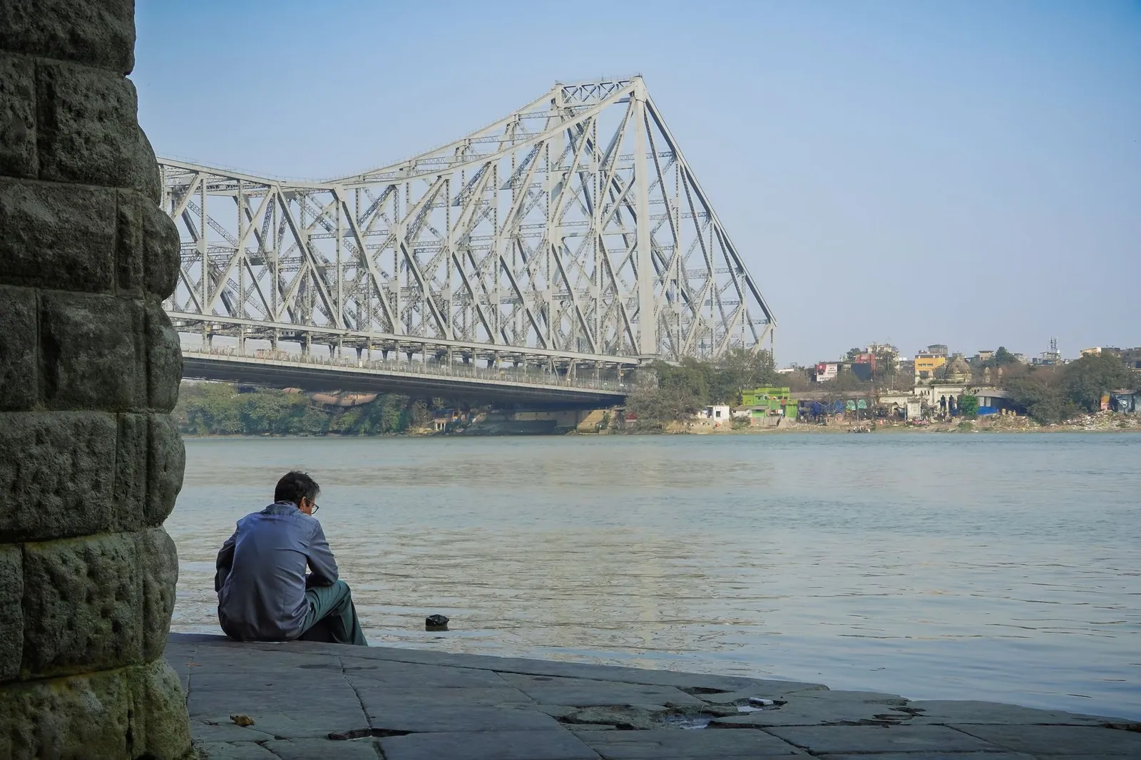 A man sits by the Hooghly River near the iconic Howrah Bridge in Kolkata, India, reflecting in solitude.