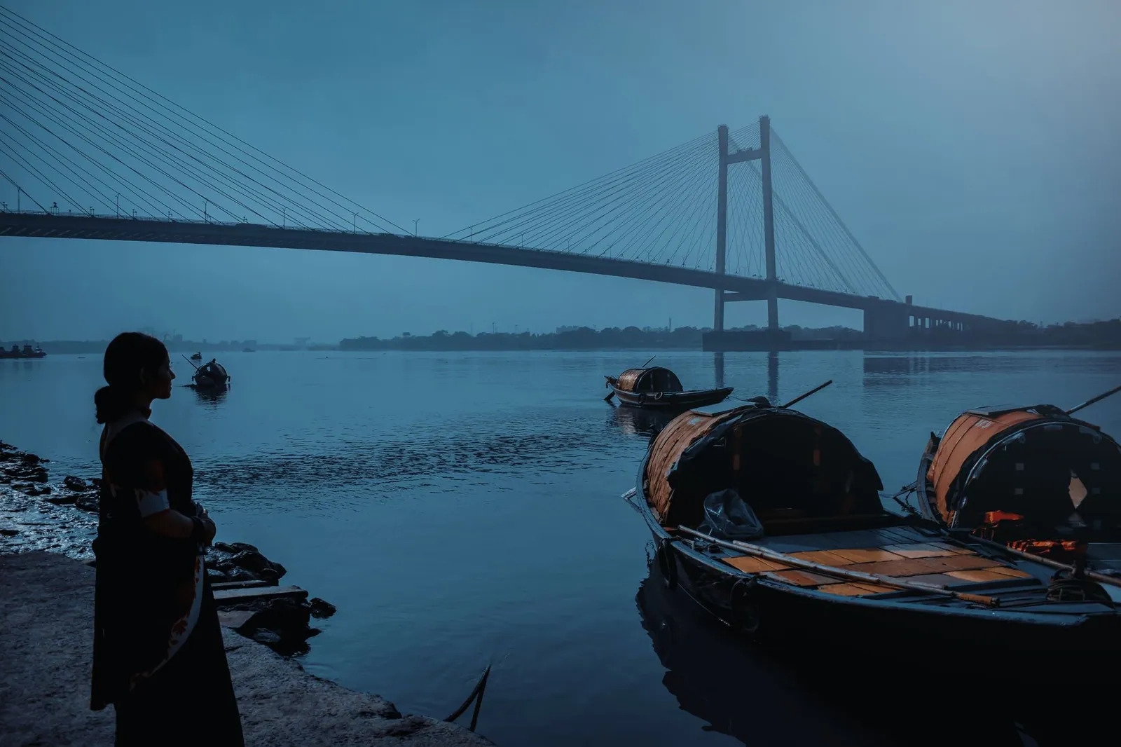 A woman by boats near the Howrah Bridge at dawn in Kolkata, India