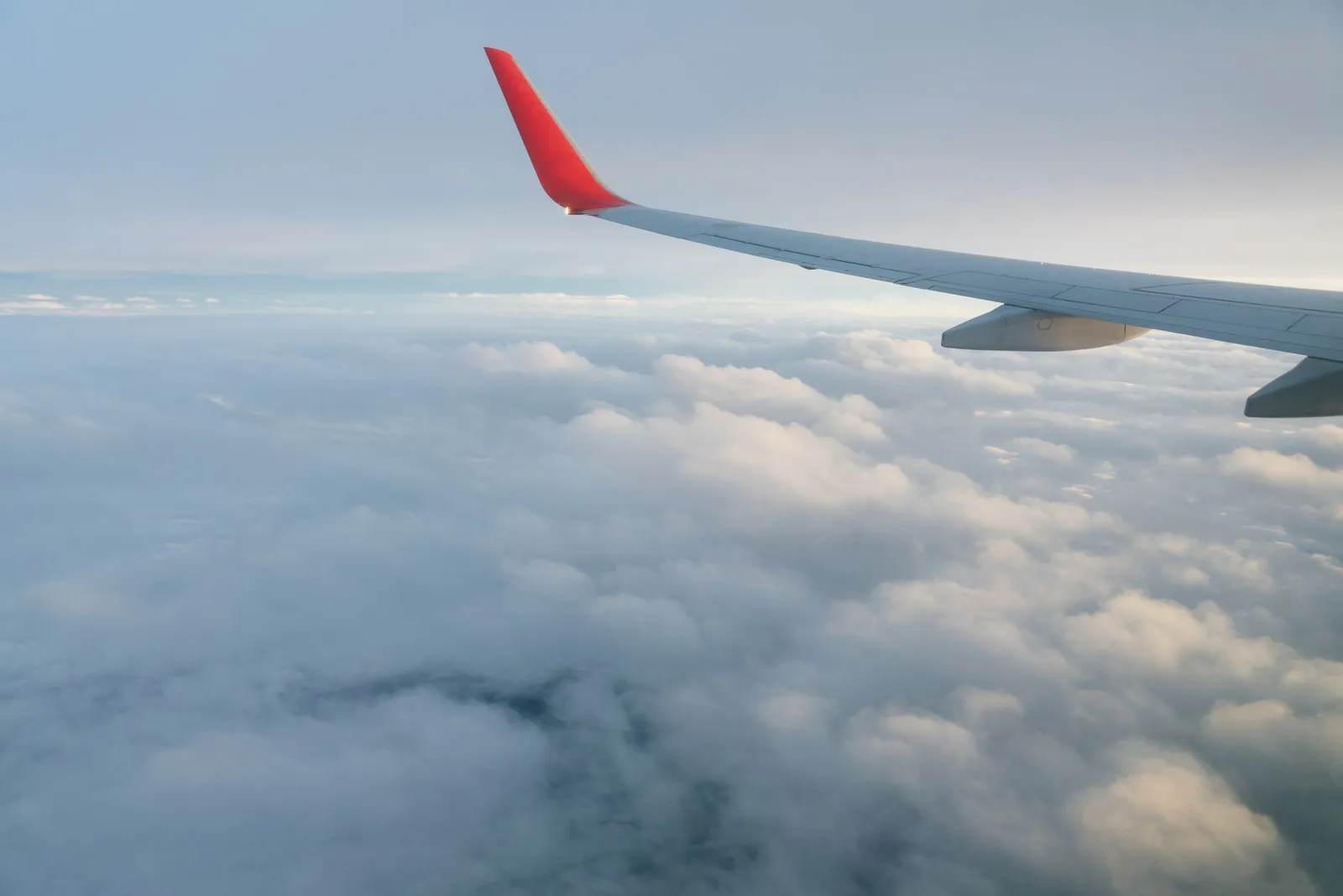 Through the window, the red and white wing of an aircraft flying over clouds in a blue sky